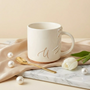 A white ceramic mug with a gold letter 'U' and gold trim sits on a marble surface. It is surrounded by a beige silk cloth, a gold spoon, white pearls, and a white tulip.