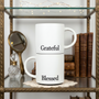 Two white ceramic coffee mugs featuring 'Blessed' and 'Grateful' in black lettering, filled with latte art, set on a marble kitchen counter in a cozy home lifestyle setting.