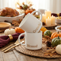 A festive Thanksgiving table setting featuring stacked 'Blessed' and 'Grateful' gold-lettered coffee mugs, surrounded by pumpkins, autumn harvest decor, and candles.